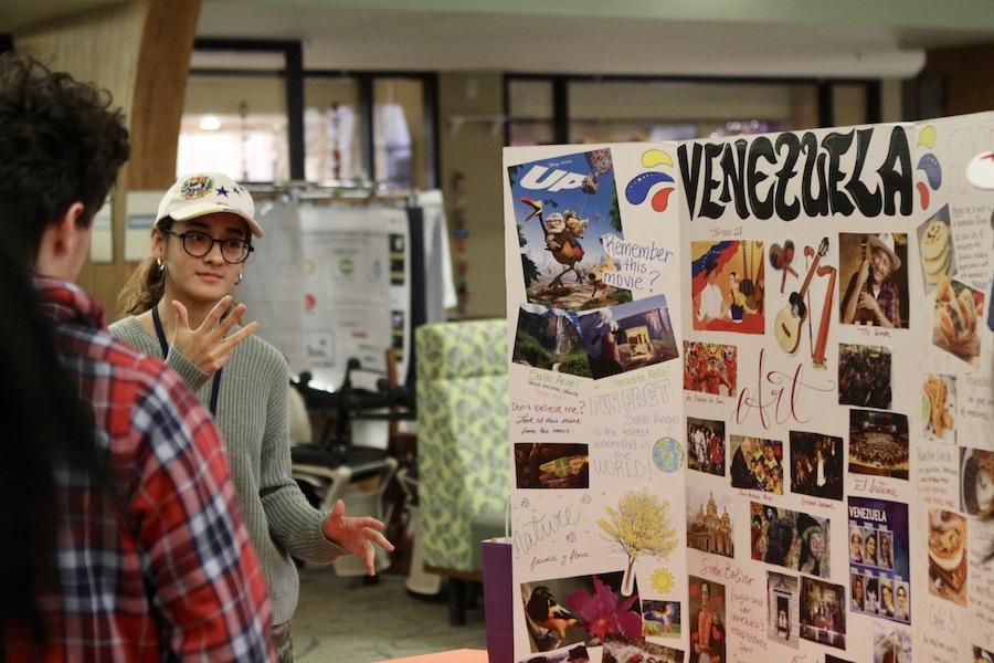 A girl with glasses talks to a boy in a red plaid shirt about a large white posterboard, covered with pictures and with the title "Venezuela." 